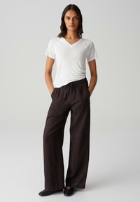 Weißes V-Ausschnitt-T-Shirt mit kurzen Ärmeln, kombiniert mit lockeren, dunkelbraunen, elastischen Hosen mit Gummizug. Das Model steht mit den Händen in den Taschen.