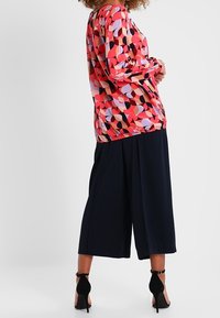 Femme portant un haut à manches longues aux motifs géométriques colorés, un pantalon large noir coupé au-dessus de la cheville, et des chaussures noires à talons avec bride à la cheville.