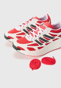 Scarpe da ginnastica rosse e bianche con tomaia in mesh, accenti neri, design a fessure e lacci in tessuto. Include due paia aggiuntivi di lacci rossi.