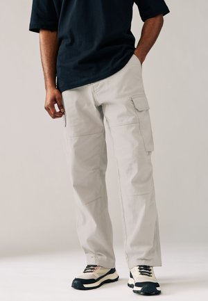 Mann trägt hellgraue Cargo-Hosen mit Seitentaschen, ein schwarzes Shirt und weiße und schwarze Turnschuhe, steht mit einer Hand in der Tasche.