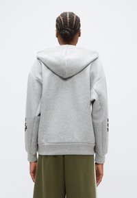 Γκρι φούτερ με κουκούλα, χαλαρή εφαρμογή, ριμπ μανσέτες και λογότυπο στο αριστερό μανίκι, σε συνδυασμό με χακί παντελόνι.