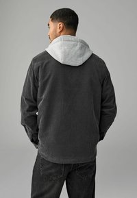 Herre corduroy jakke i mørkegrå, med grå hætte og ribbet tekstur. Designet inkluderer en afslappet pasform og skjulte knapdetaljer.
