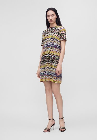 Femme debout portant une robe à manches courtes à motif zigzag multicolore et des sandales noires à talons hauts sur un fond gris uni.