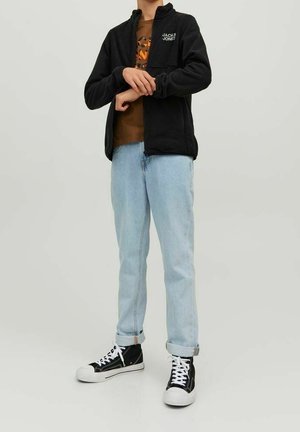 Personne portant une veste noire Jack & Jones, un t-shirt graphique marron, un jean bleu clair retroussé aux chevilles et des baskets montantes noires à lacets blancs.