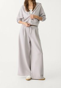 Hellgrauer, kürzerer Hoodie mit Kordelzug, aus weichem Material, und zwei vorderen Taschen, kombiniert mit weit geschnittenen Sweatpants und weißen Sneakern.
