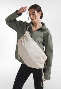 Leichte olivgrüne Jacke über einem engen schwarzen Top; eine beige Umhängetasche mit Zugbandverschluss und Logo, die eine glatte, matte Textur zeigt.