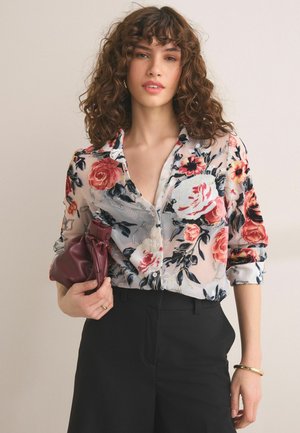 Femme aux cheveux bouclés portant un chemisier transparent à fleurs et un pantalon noir, tenant une pochette en cuir bordeaux dans sa main gauche.
