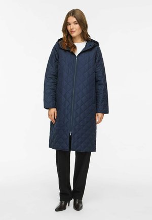 Femme aux cheveux bruns portant un long manteau matelassé bleu marine avec capuche, pantalon noir et bottes à talons noires, debout sur un fond blanc.