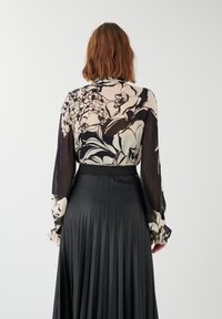Jupe plissée noire associée à un haut transparent avec un grand motif floral en crème et noir. Manches longues avec des poignets froncés.
