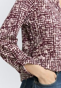 Chemise bordeaux avec un motif abstrait crème texturé. Manches longues avec des poignets à boutons, présentée rentrée dans un jean bleu. Tissu lisse, design décontracté.