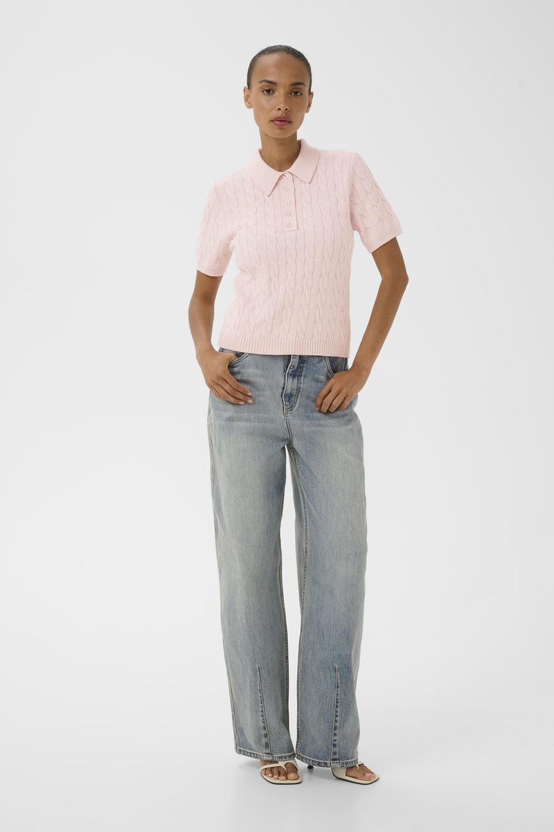 Femme debout, les mains dans les poches, portant un polo en maille rose clair à manches courtes, un jean large bleu clair et des sandales à bouts ouverts.