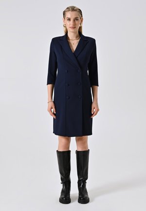 Donna che indossa un abito blazer doppiopetto blu navy con maniche a tre quarti e stivali in pelle neri al ginocchio, in piedi contro uno sfondo neutro.