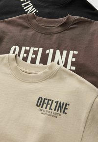 Trois sweat-shirts empilés : noir, marron et beige. Chacun présente un texte "OFFL1NE" en gras dans des couleurs contrastantes. Tissu en coton, design à col rond.