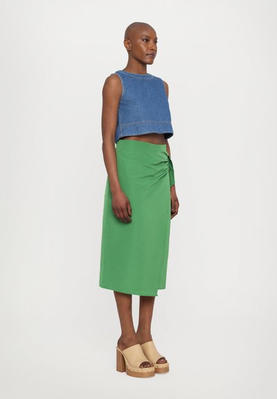 Denim uskičeni top, zeleni omotani suknja, smeđi drveni klomfi, glatki materijal, minimalistički dizajn, model stoji na jednostavnoj pozadini.