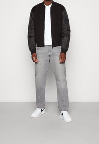 Zwarte bomberjack met gewatteerde mouwen, wit T-shirt, lichtgrijze jeans en witte sneakers met zwarte accenten, leunend tegen een neutrale achtergrond.