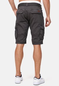 Homme portant un short cargo noir avec poches à rabat et des baskets blanches Reebok, debout dos au spectateur sur un fond uni.