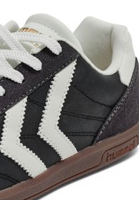 Baskets en cuir noir et blanc avec lacets blancs, semelle marron, col texturé et logo "hummel" en relief sur le côté.