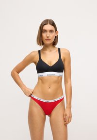Bralet negru cu bandă albă lată care prezintă logo-ul "Calvin Klein", asortat cu un thong roșu cu bandă albă corespunzătoare. Material fin, design minimalist.