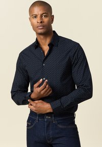 Camisa de botones azul marino con un pequeño patrón de puntos blancos, manga larga y cuello clásico, combinada con jeans de mezclilla oscura.