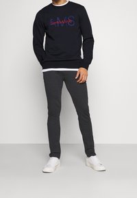 Námořnická crewneck mikina s vyšíváním "Scotch&Soda" a "AMS" v červené a modré barvě, v kombinaci s tmavě šedými slim-fit kalhotami a bílými teniskami.