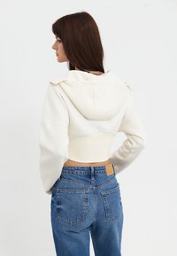 Zugeschnittenes weißes Sweatshirt mit Kapuze und geripptem Saum, getragen mit blauen hochgeschnittenen Jeans, die ein Lederpatch und zwei Hintertaschen haben.