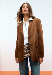 Blazer en daim marron avec deux boutons, revers à cran et poches avant, porté sur un haut zippé blanc et une jupe à motifs. Coupe classique, tissu texturé.