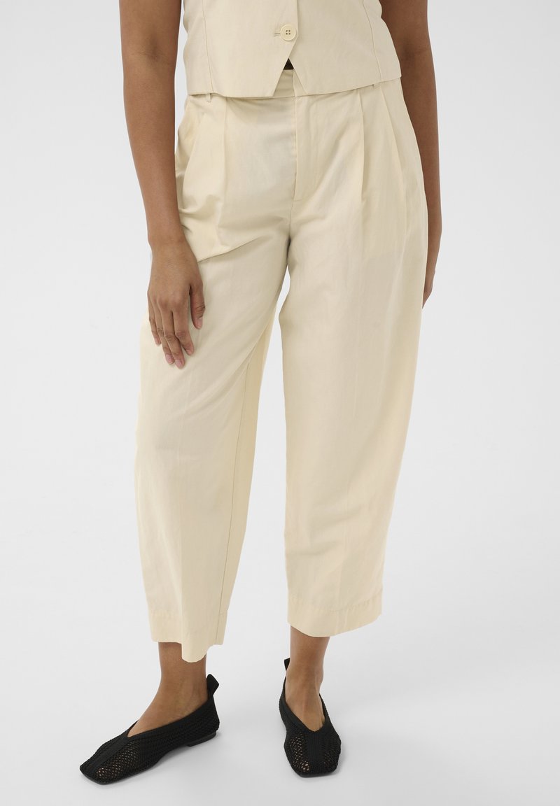 Femme portant un pantalon beige taille haute raccourci avec des plis, un gilet beige assorti, et des chaussures plates tissées noires, debout sur un fond blanc.