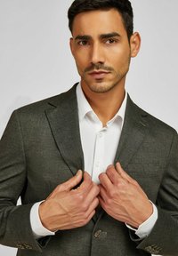 Grau strukturierter Blazer mit Revers, Ein-Knopf-Verschluss und einer Vorderbrusttasche, getragen über einem weißen Hemd mit Knöpfen.