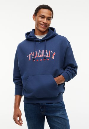 Jeune homme souriant portant un sweat à capuche bleu marine avec le logo "Tommy Jeans" et un jean bleu, debout sur un fond blanc.