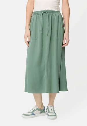 Persoon draagt een hooggetailleerde midi-rok in saliegroen met trekkoord en opgestikte zakken, gecombineerd met witte sneakers met groene en blauwe accenten.