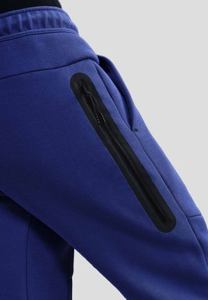 Pantalones deportivos azules con un bolsillo con cremallera negra en el muslo, textura de tela suave y cinturilla elástica.