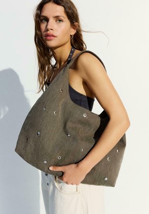 Nainen, jolla on laineikkaat ruskeat hiukset, pukeutuneena hihattomaan mustaan toppiin ja valkoisiin housuihin, pitää kädessään suurta ruskeaa kangaskangasta metallirenkailla.