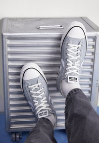 Une personne portant des baskets Converse All Star grises claires et des chaussettes blanches repose ses pieds sur une valise rigide argentée à roulettes.