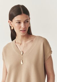 Beige gebreide trui met een V-hals en korte mouwen, gecombineerd met gouden creolen en een gouden hangersnoer. Zachte textuur.