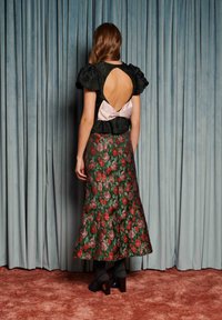 Žena stojí čelom k modrým závesom, má na sebe čierny top s nafúknutými rukávmi a otvoreným chrbtom s ružovou mašľou, kvetovanú midi sukňu a tmavé lodičky na podpätku.