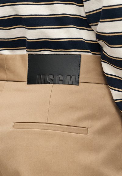 Μπεζ παντελόνι με πίσω τσεπάκι welt και μαύρο ορθογώνιο έμπλαστρο με ανάγλυφη επιγραφή «MSGM», σε συνδυασμό με λευκό, ναυτικό και καφέ ριγέ πουκάμισο.