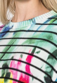 Gros plan sur une personne portant un pull rayé coloré avec des lignes horizontales vertes, bleues, roses et noires, et un col côtelé.