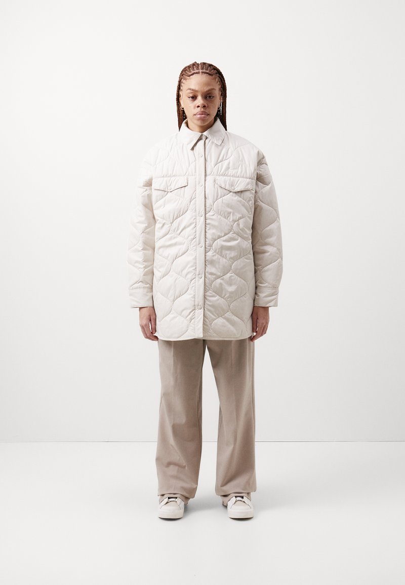 Jachetă cu țesătură bej deschis, matlasată, cu guler, având două buzunare pe piept și o croială largă, asociată cu pantaloni bej cu picior larg și adidași.