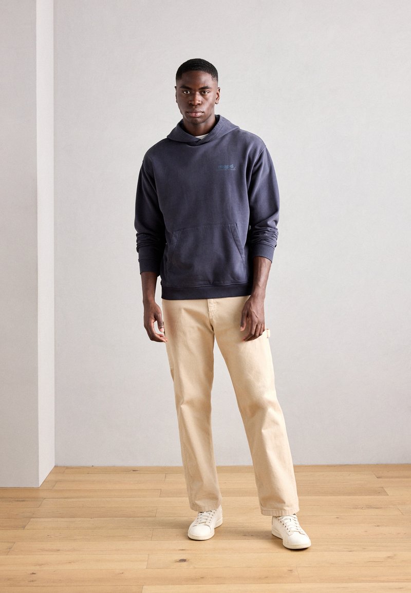 Marineblaue Kapuzenjacke mit einer Fronttasche und Kordelzug, kombiniert mit beigefarbenen Hosen und weißen Sneakers, vor einem neutralen Hintergrund.