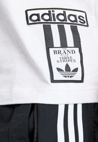 Baltas medvilnės audinys, kuriame įsiūtas "adidas" logotipas, juodas pleistras su užrašu "PREKĖ SU TRIMIS JUOSTOMIS" ir trys vertikalios juodos juostos.
