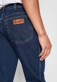 Nahaufnahme einer Person, die dunkelblaue Wrangler-Jeans trägt, wobei die hintere Tasche mit dem Lederschild der Marke und dem gestickten "W"-Design zu sehen ist.
