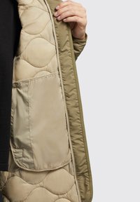 Padded jacket s khaki exteriérom, béžovou podšívkou, prešívaným vzorom a vreckom na klopu. Interiér má hladkú textúru.
