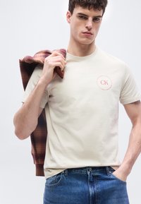 Νεαρός άνδρας που φοράει κρεμ t-shirt CK και μπλε τζιν, κρατώντας ένα καρό καφέ σακάκι στον ώμο του, μπροστά σε έναν απλό φόντο.