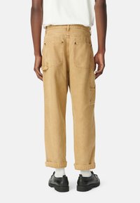 Pantalones cargo de pierna ancha color beige, fabricados en algodón, con costuras visibles, dos bolsillos traseros, dobladillo recogido y combinados con zapatos negros y calcetines blancos.