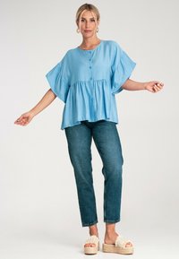 Jasnoniebieska bluzka z krótkim rękawem i zapinaniem na guziki, luźny krój oraz falbaniasty dół. Połączona z ciemnoniebieskimi dżinsami i beżowymi sandałami na platformie.