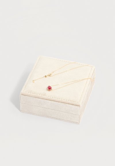 Λεπτό χρυσό κολιέ με μικρό κόκκινο μενταγιόν σε σχήμα καρδιάς, τοποθετημένο πάνω σε βελούδινο κουτί κοσμημάτων εκρού χρώματος.
