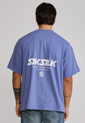 Uomo con capelli scuri che indossa una t-shirt blu SikSilk con testo bianco sul retro, abbinata a jeans blu, in piedi di spalle.