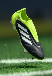 Green and black soccer shoe with a textured upper, three white stripes, and spike studs, hovering above grass with small debris.