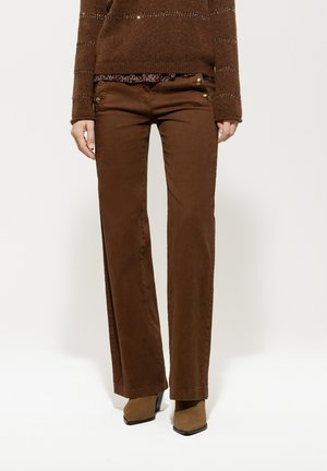 Braune, gerade geschnittene Hose mit glatter Textur und dekorativer goldfarbener Hardware an den Taschen. Kombiniert mit einem braunen Strickpullover.