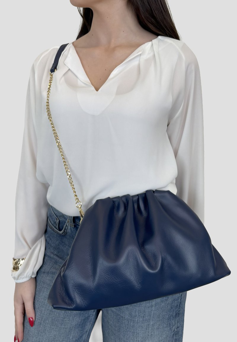 Mujer con blusa blanca y jeans lleva un gran bolso de cuero azul marino con cadena dorada al hombro.